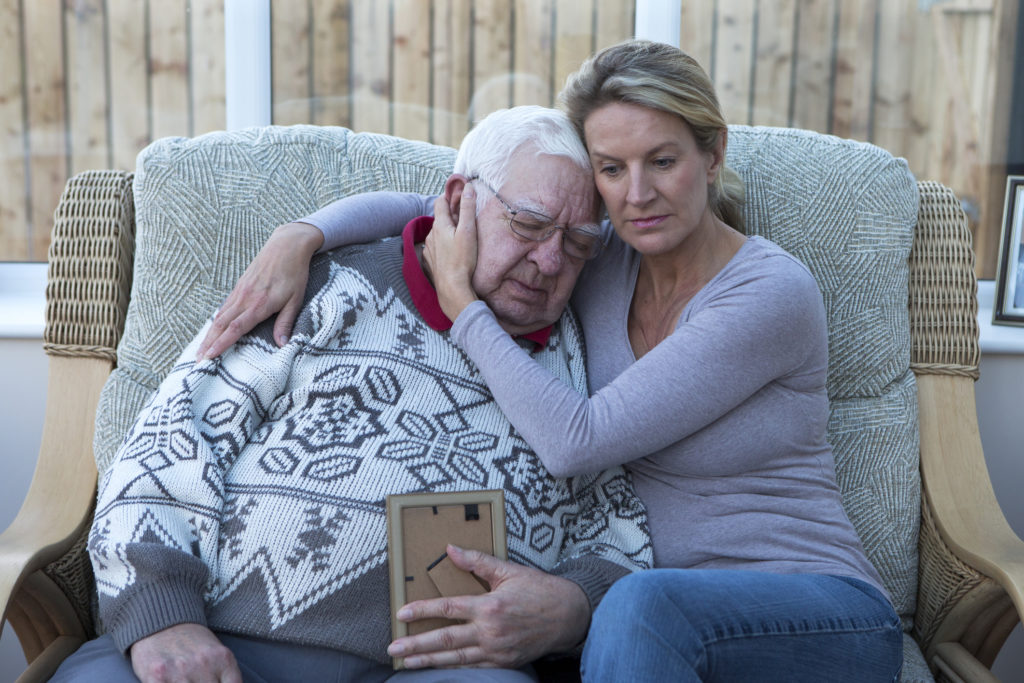 A shot of a senior man sitting on the sofa with his daughter, looking at a photograph. He is cuddling his daughter, looking sad and holding the photo.