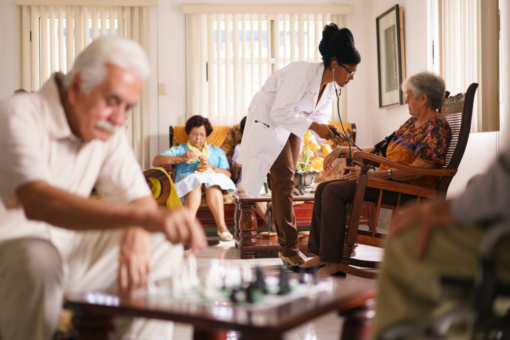 Old people in geriatric hospice: Black doctor visiting an aged patient, measuring blood pressure of a senior woman. Group of retired men in foreground playing chess.