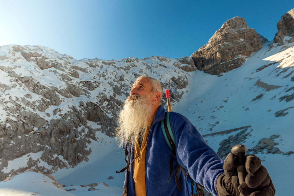 Happy senior man with long white beard is climbing on the snowy Kanin, Julian Alps, Slovenia-Italy border, Primorska region, Europe. He is wearing sunglasses, a backpack with an ice ax. Sun behind him, blue clear sky, copy space. Nikon D3x