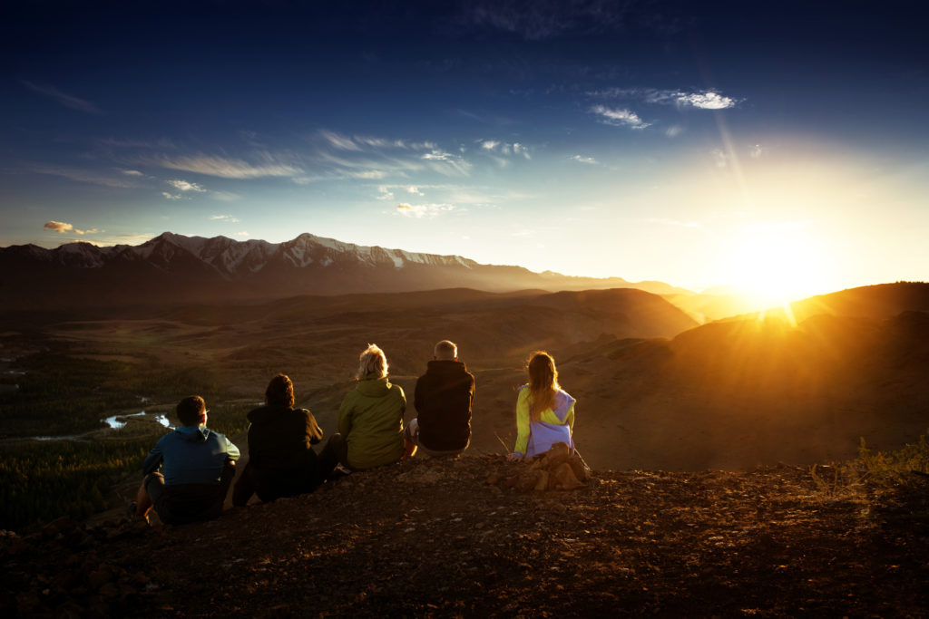 Group of friends sits on the mountain's top on background of mountains and sunset