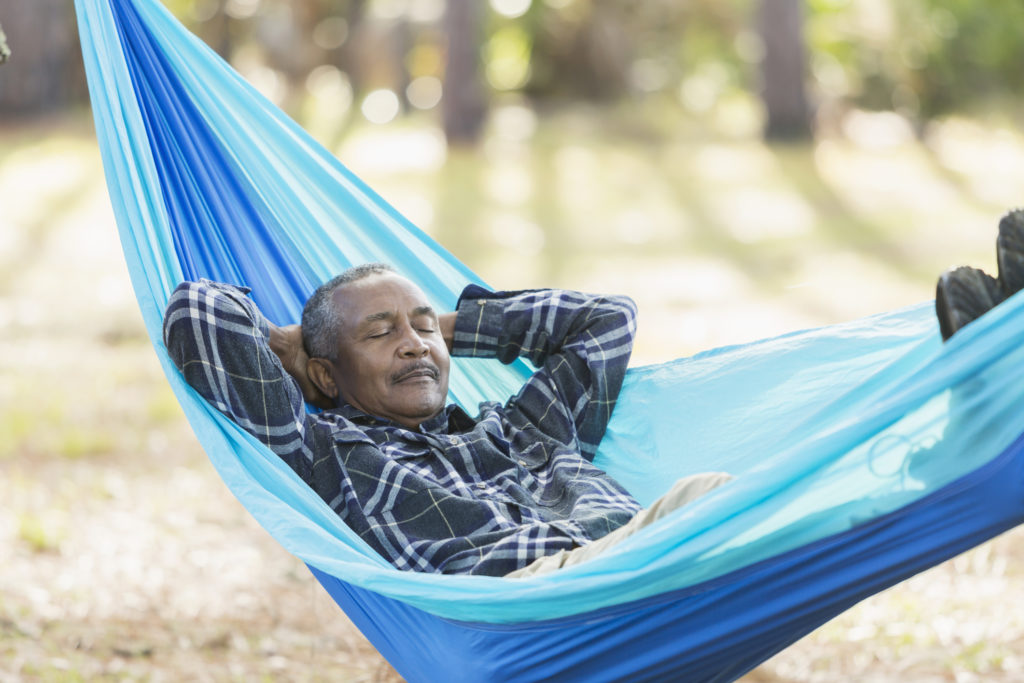 A senior African American man sound asleep in a hammock in the park. His eyes are closed, and his hands are behind his head.