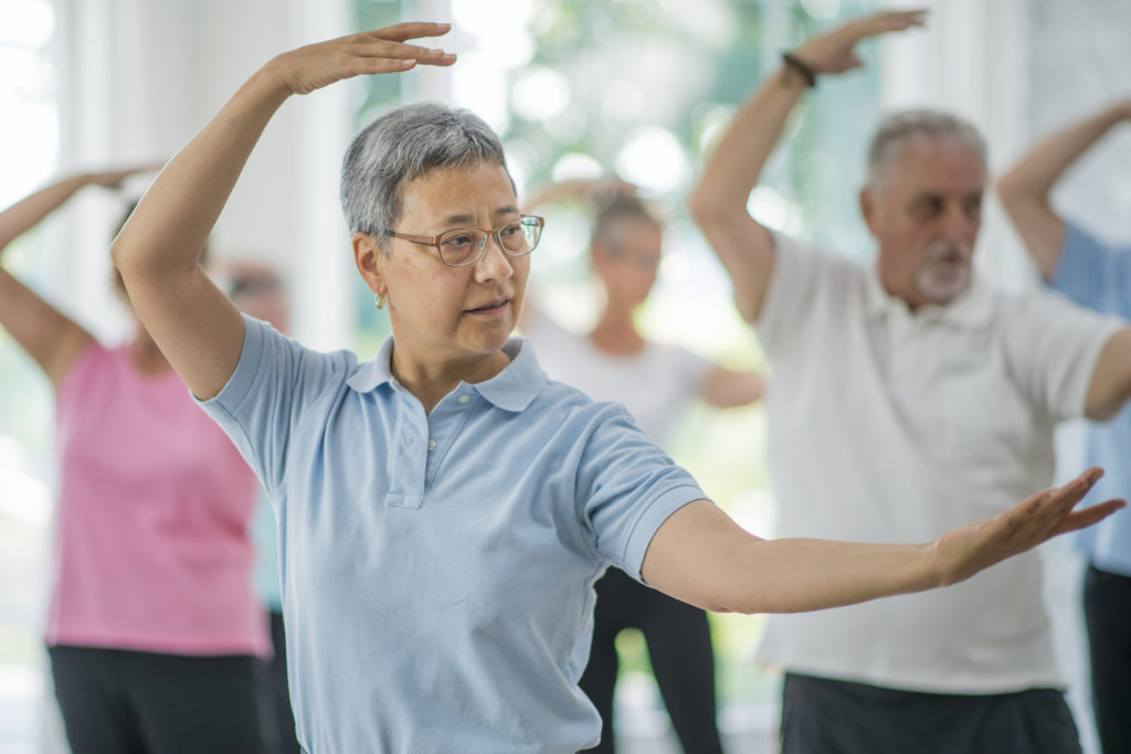A multi-ethnic group of senior adults are taking a tai chi fitness class. They are practicing their martial art inside of a brightly lit studio. Here, a woman of asian ethnicity is leading the class through movements.