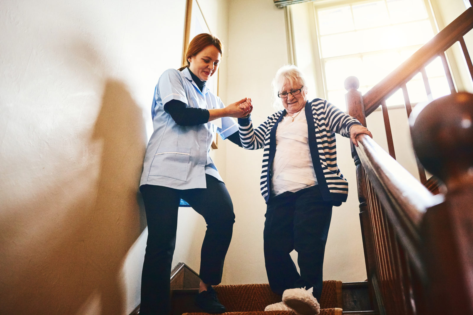 Female caregiver helping senior woman walking down stairs at home
