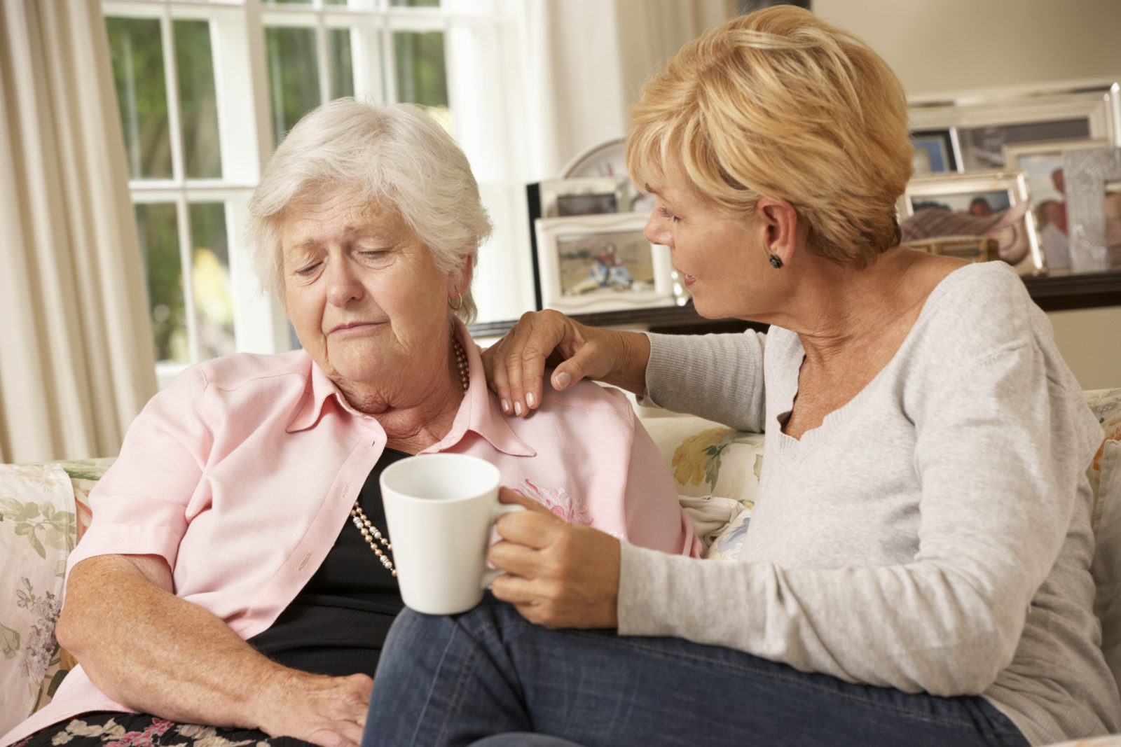 Adult Daughter Visiting Unhappy Senior Mother Sitting On Sofa At Home Trying To Comfort Her