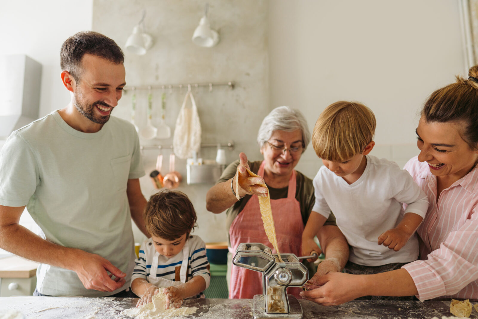 Photo of a smiling senior woman teaching her daughter, her son-in-law, and grandsons how to make homemade pasta in the kitchen; having a great time while making food and enjoying each other's company.